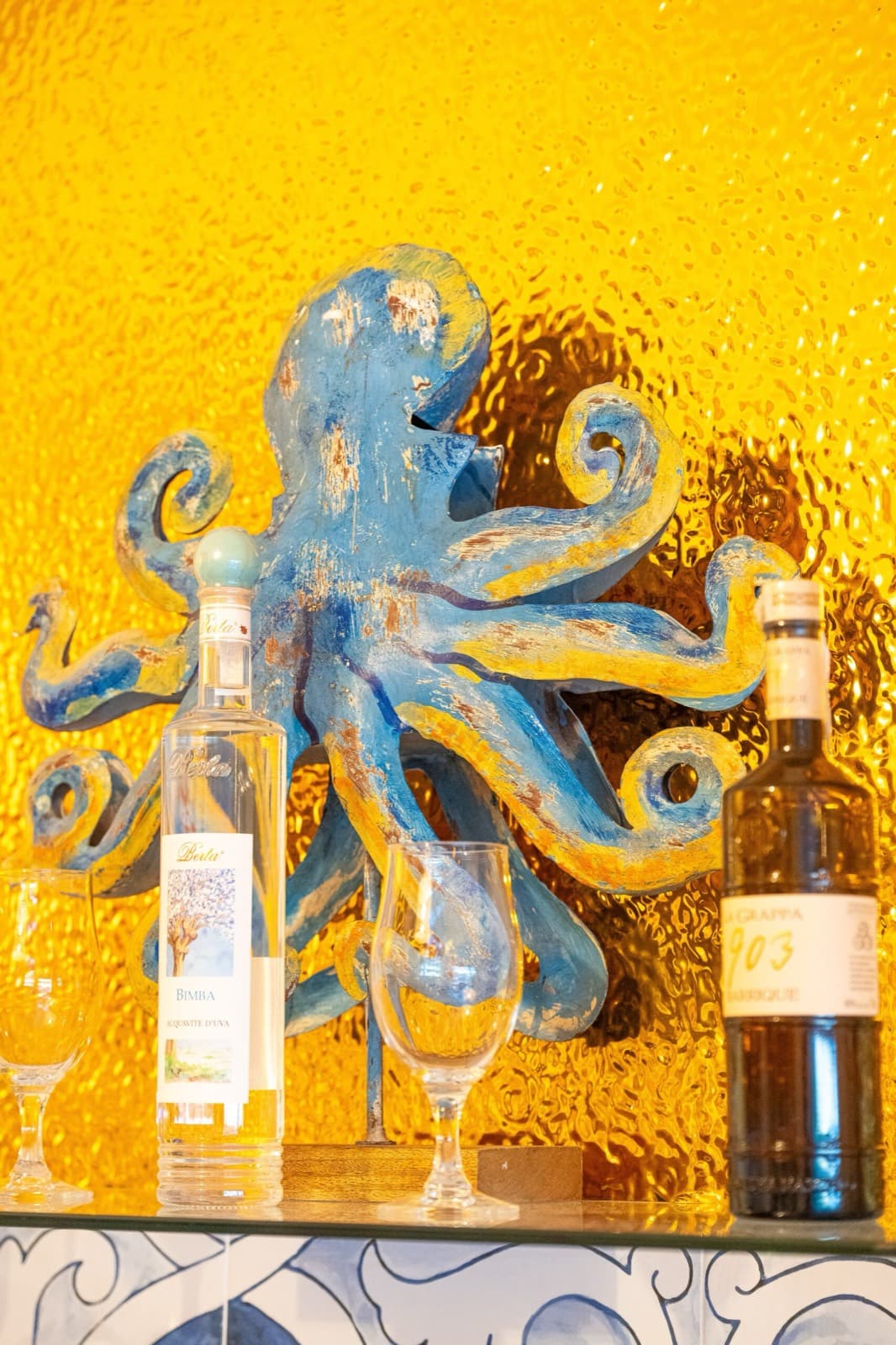 Scultura di polpo blu davanti al vetro martellato dorato, tra bottiglie di grappa